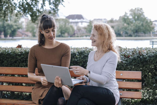 Zwei Frauen sitzen auf einer Bank am Main und tauschen sich vor einem offenen Laptop aus.
