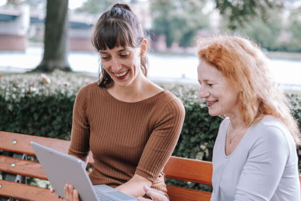 Zwei Frauen sitzen auf einer Bank am Main und tauschen sich vor einem offenen Laptop aus.